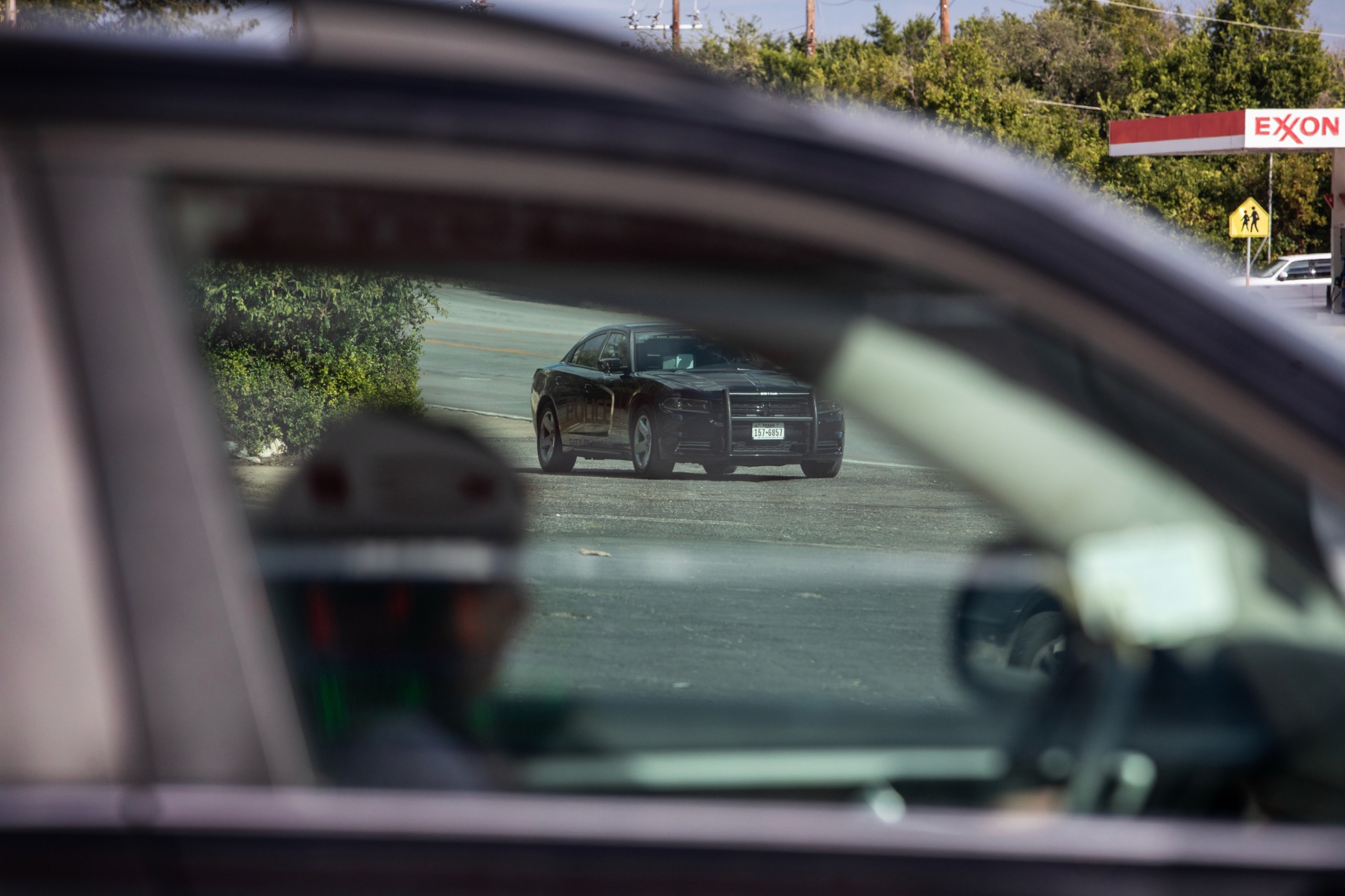 A soft-focus close-up of a person in the driver’s seat of a vehicle, viewed through the passenger window, with the black Charger clearly visible in the background and parked on the roadside.