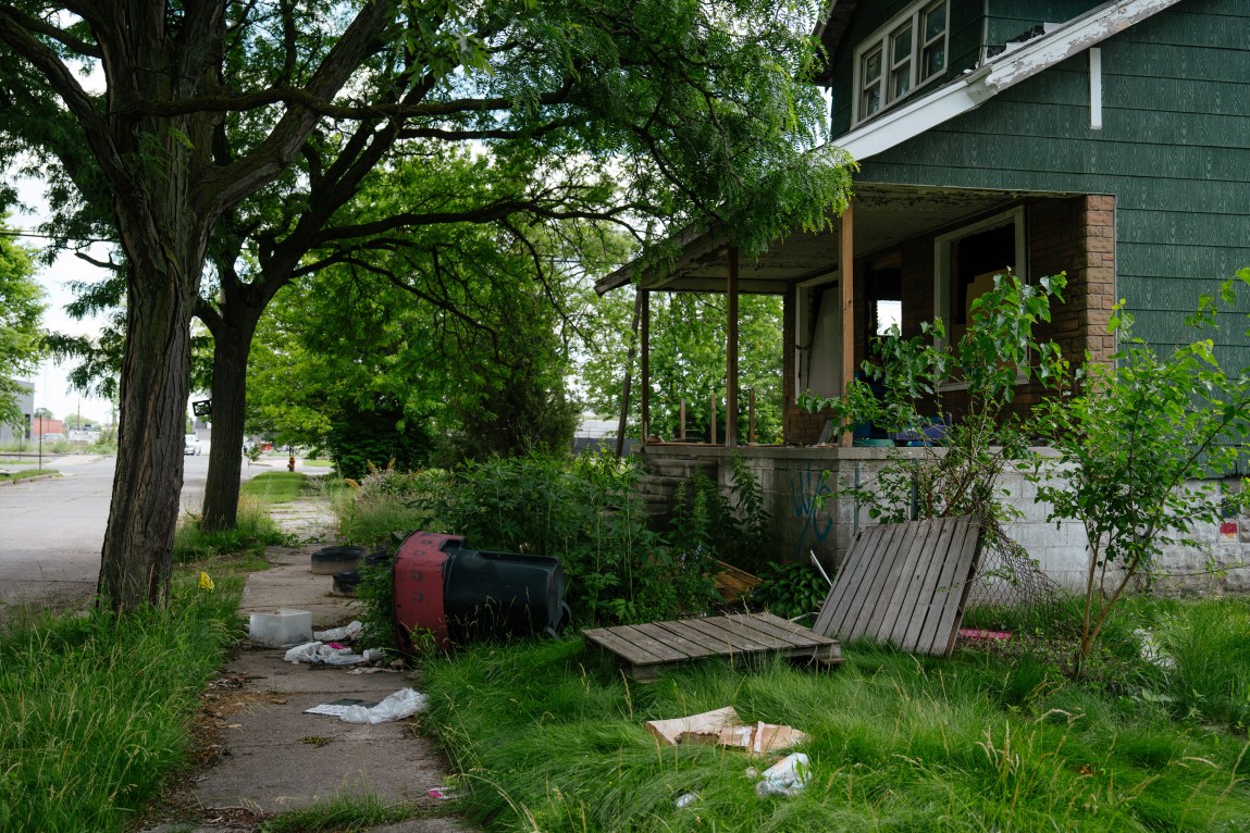 A dilapidated house with no front windows and an overgrown, trash-covered lawn.