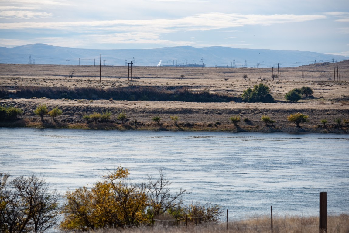Telephone poles and some low buildings stick up from the desert, as seen from across the Columbia River. Mountains rise up behind the site.