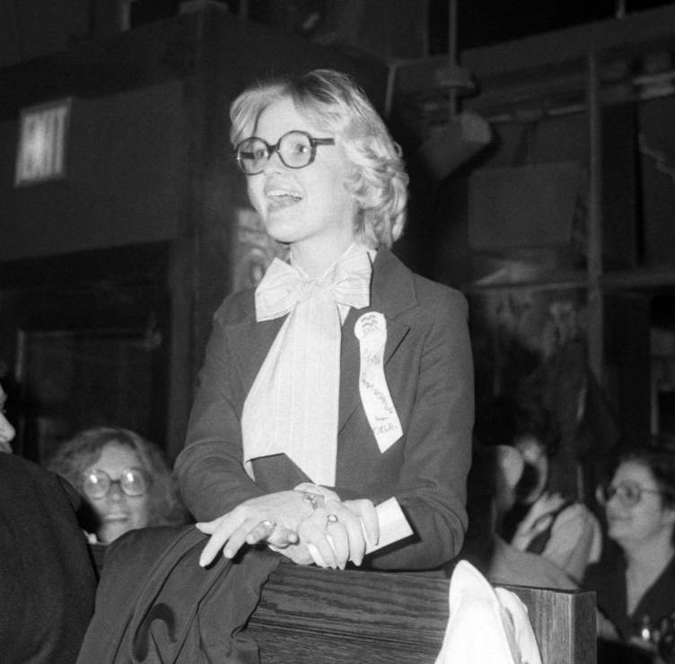 A black-and-white photograph shows a young woman smiling while wearing round glasses, a blazer, a white blouse with a large bow and a ribbon pinned to her lapel.
