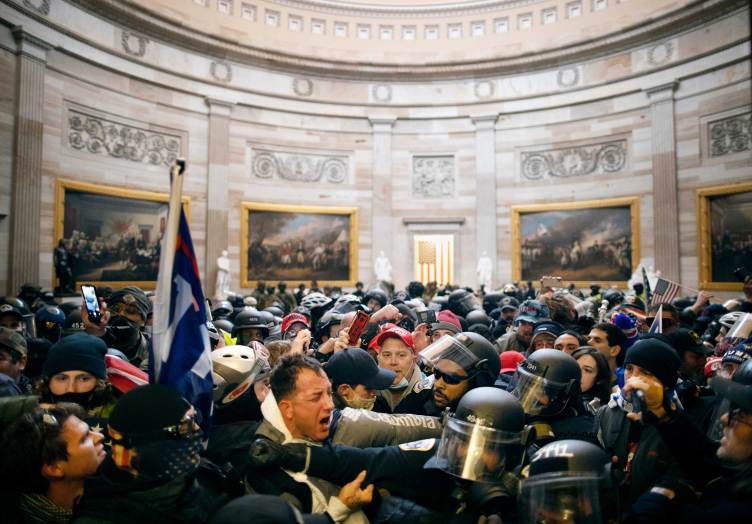 A large crowd of rioters fights with police officers wearing helmets and face shields inside the rotunda of the U.S. Capitol. Historic paintings line the curved stone walls in the background.