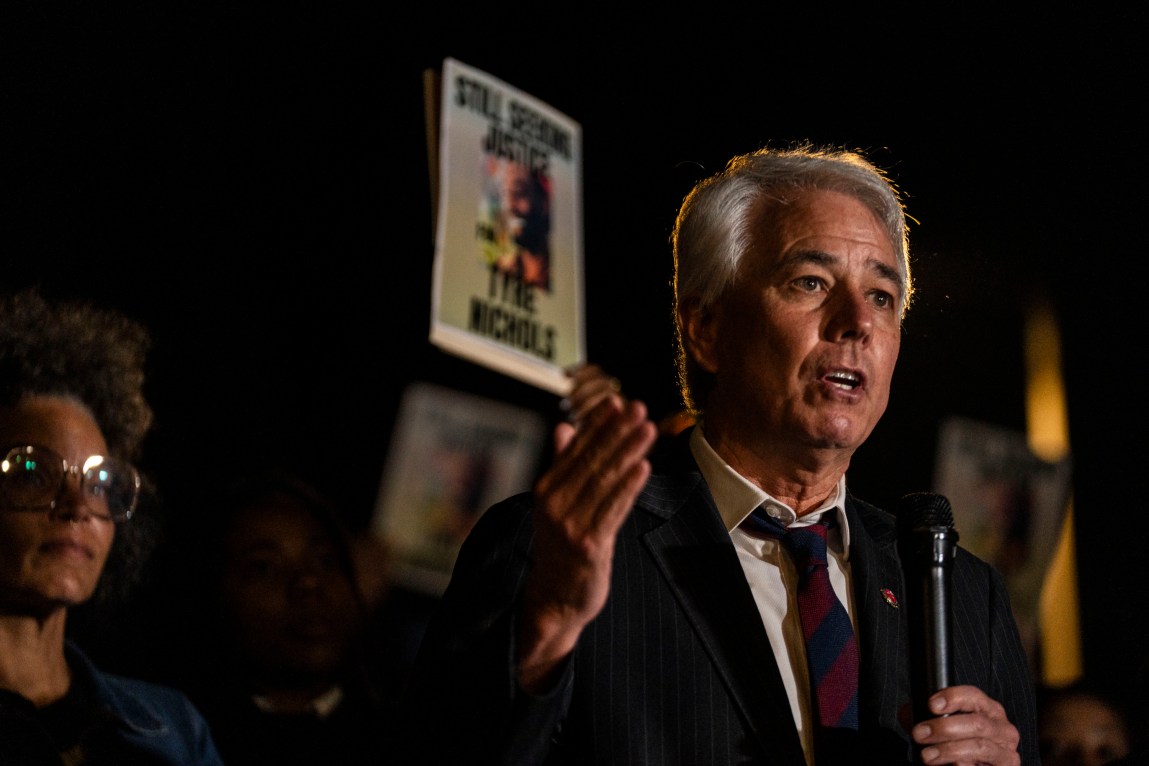 A man holding a microphone gestures with his hand as he speaks. In the background, a crowd is holding signs that say, “Still Seeking Justice. Tyre Nichols.”