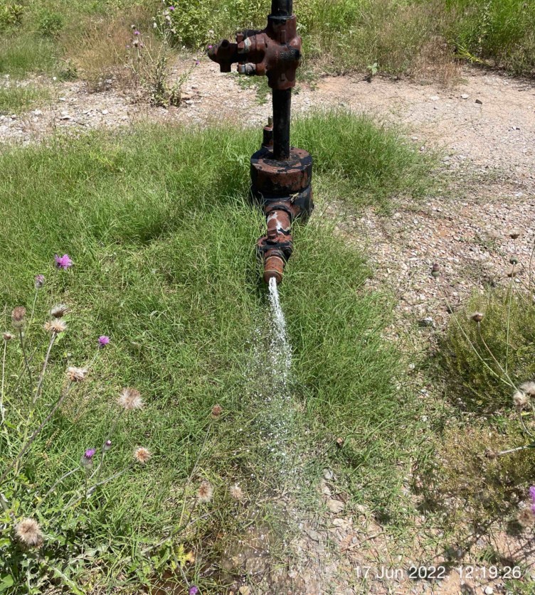 A structure of rusted pipes sprays water into an area surrounded by grass and dirt.