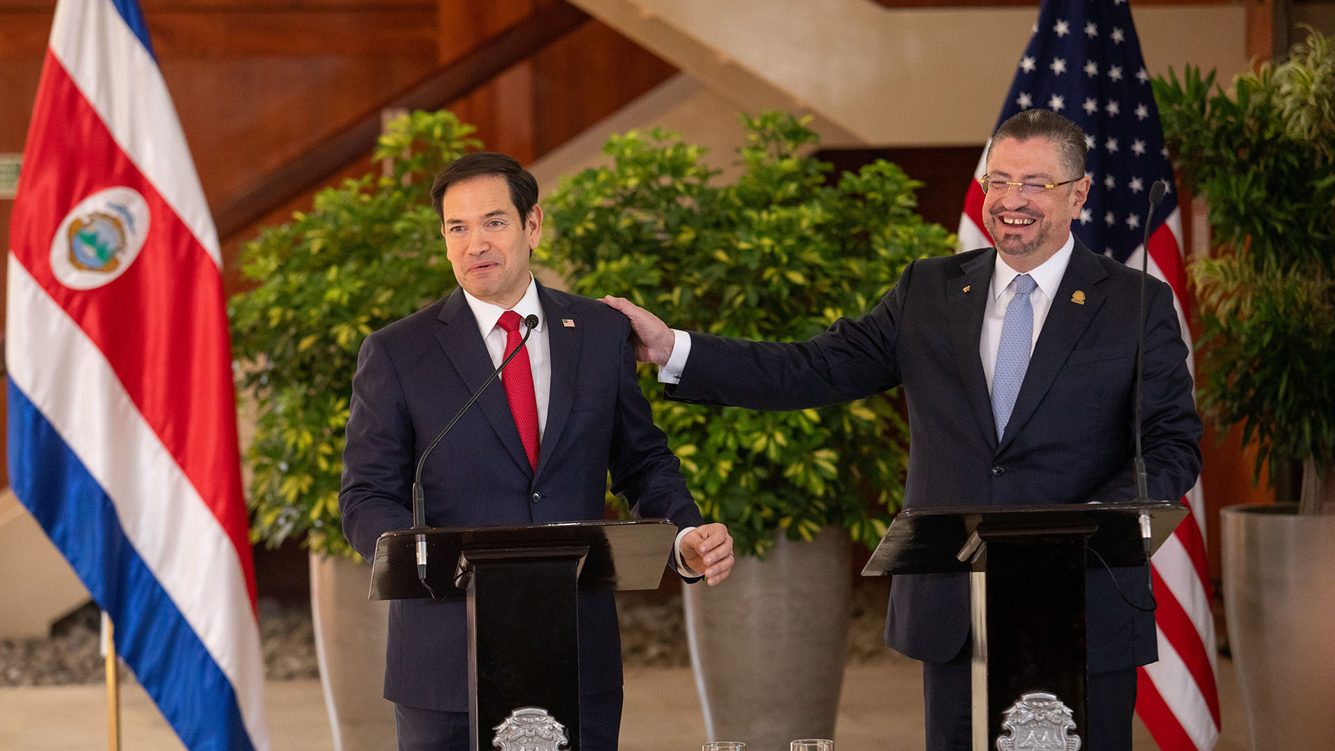 Two men in suits giving a press conference in front of Costa Rican and American flags.