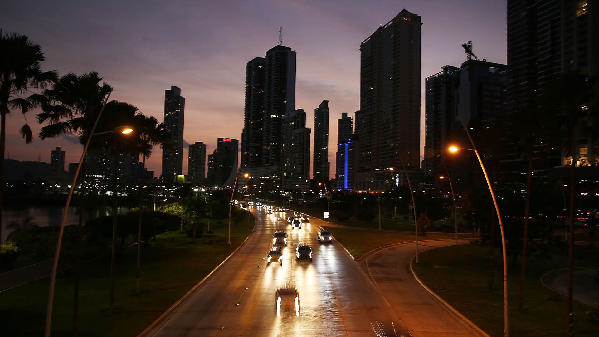 Photo of Panama City's skyline at dusk, with cars headlights illuminating a road in front of tall buildings.