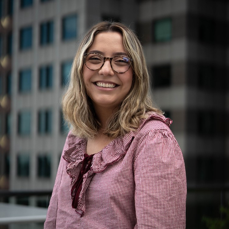 A woman with shoulder-length blonde hair smiles at the camera. She wears glasses and a red patterned blouse and is in front of out-of-focus buildings.